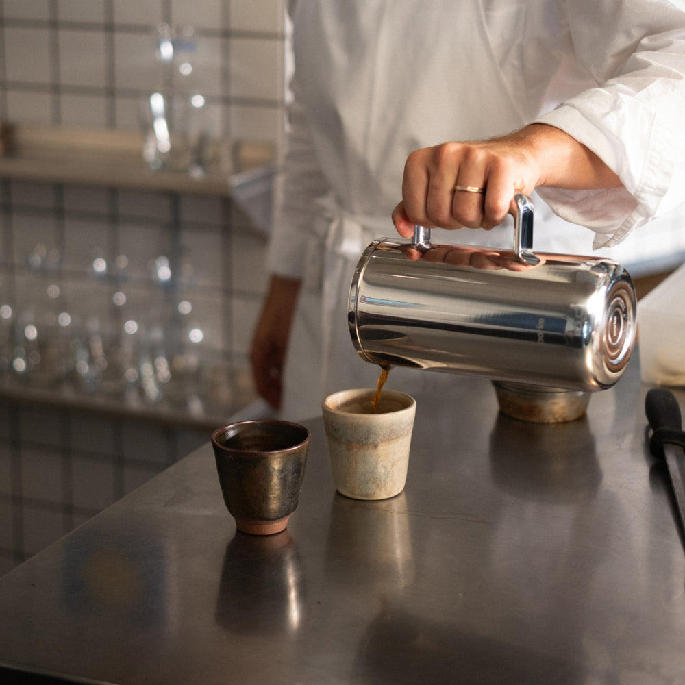 Person pouring coffee from a steel Coffee Press into a ceramic cup on a reflective surface.