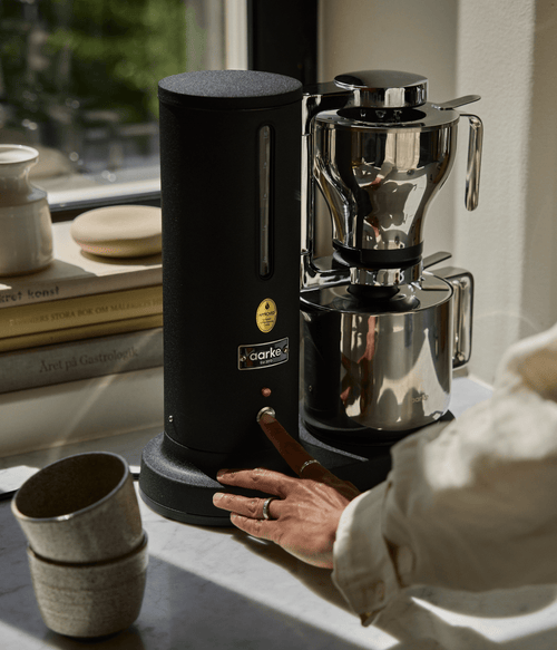 Aarke Coffee Maker in Matte Black on a counter brewing directly in Thermal Jug, surrounded by books in the background.