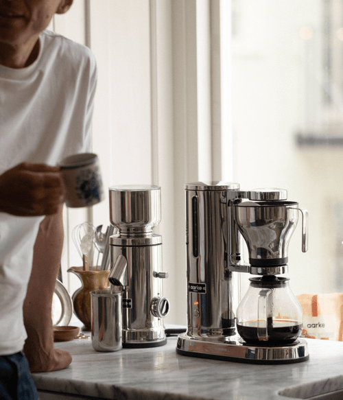 A person holding a coffee cup beside the Aarke stainless steel coffee grinder and coffee maker, with freshly brewed coffee in the glass carafe.
