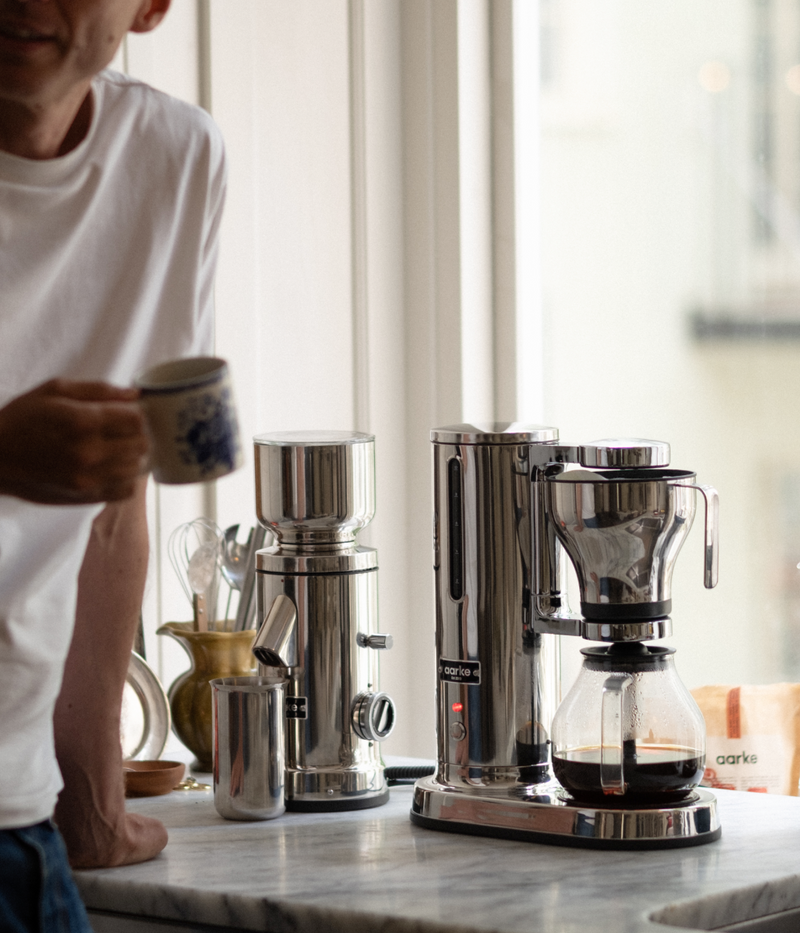 A person holding a coffee cup beside the Aarke stainless steel coffee grinder and coffee maker, with freshly brewed coffee in the glass carafe.